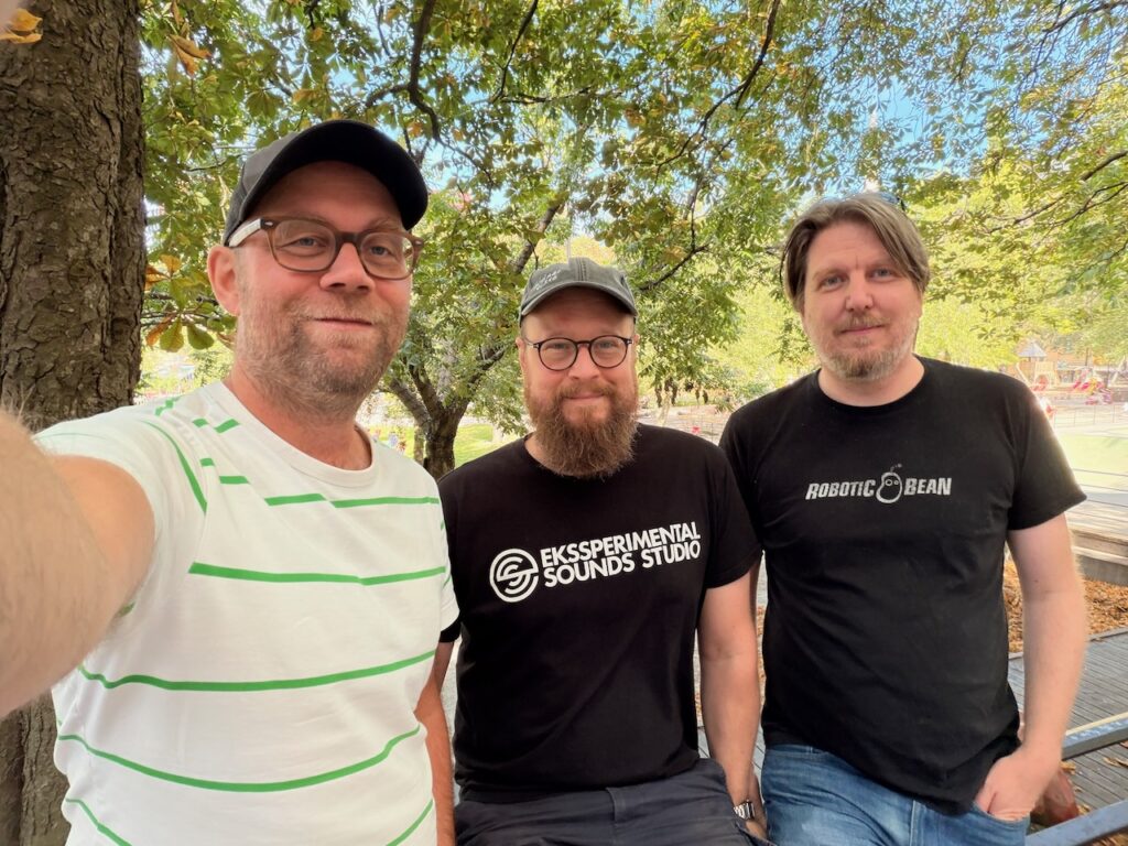 Three men in a selfie against green trees in a park.